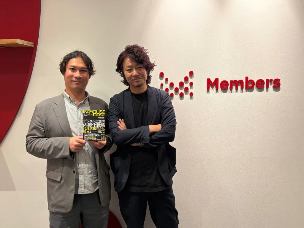 壁面のメンバーズロゴの前で並び立つ仲山氏（写真左）と田中（写真右）